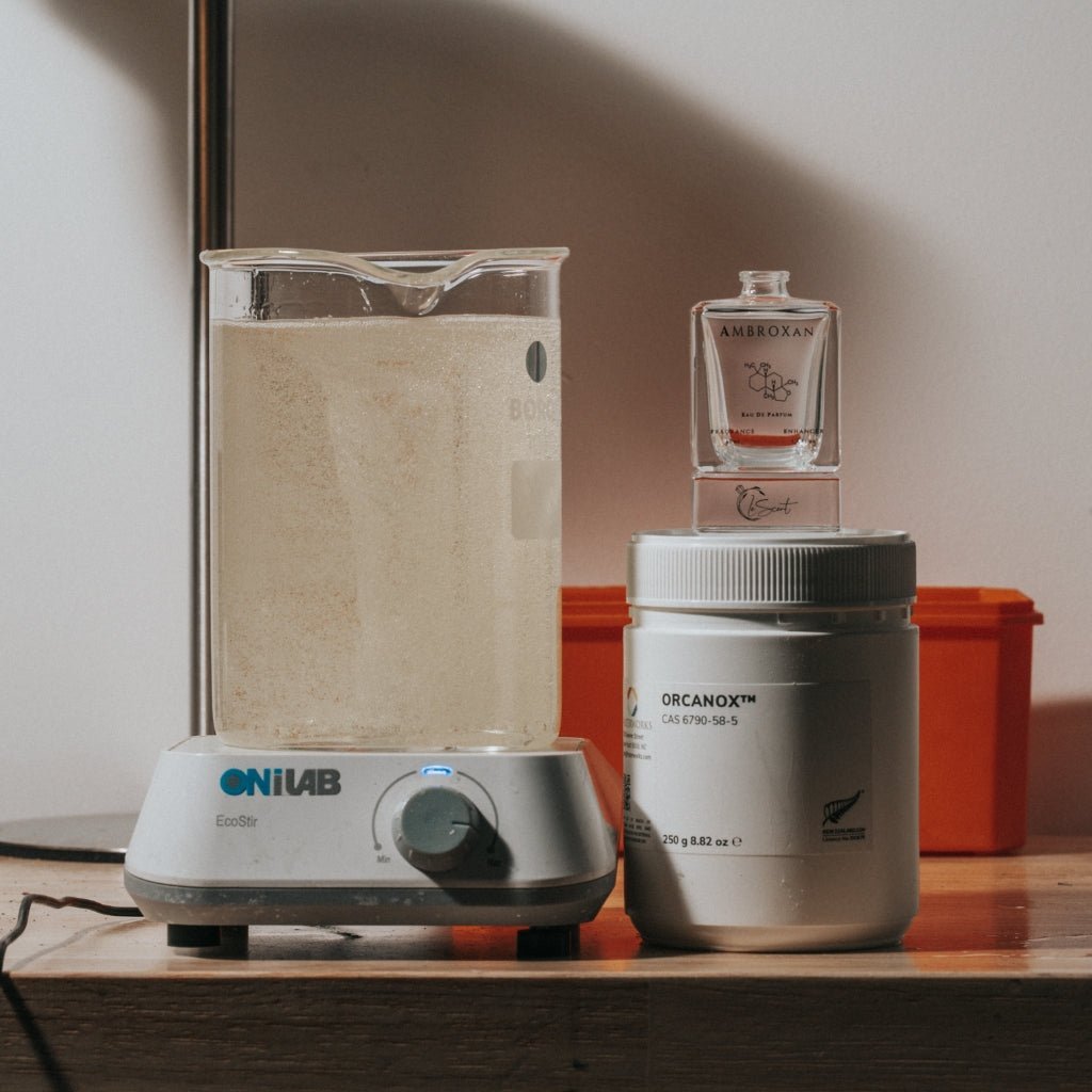 LeScent Ambroxan perfume bottle and Orcanox compound beside a mixing beaker in a fragrance lab, representing small-batch formulation and ingredient blending.
