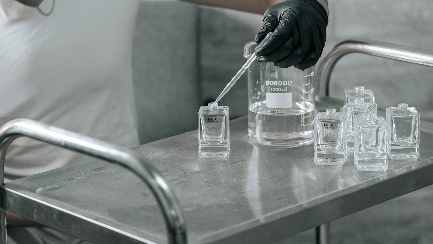 A person wearing black gloves carefully uses a glass pipette to fill a small, square LeScent perfume bottle from a large beaker on a stainless steel cart.