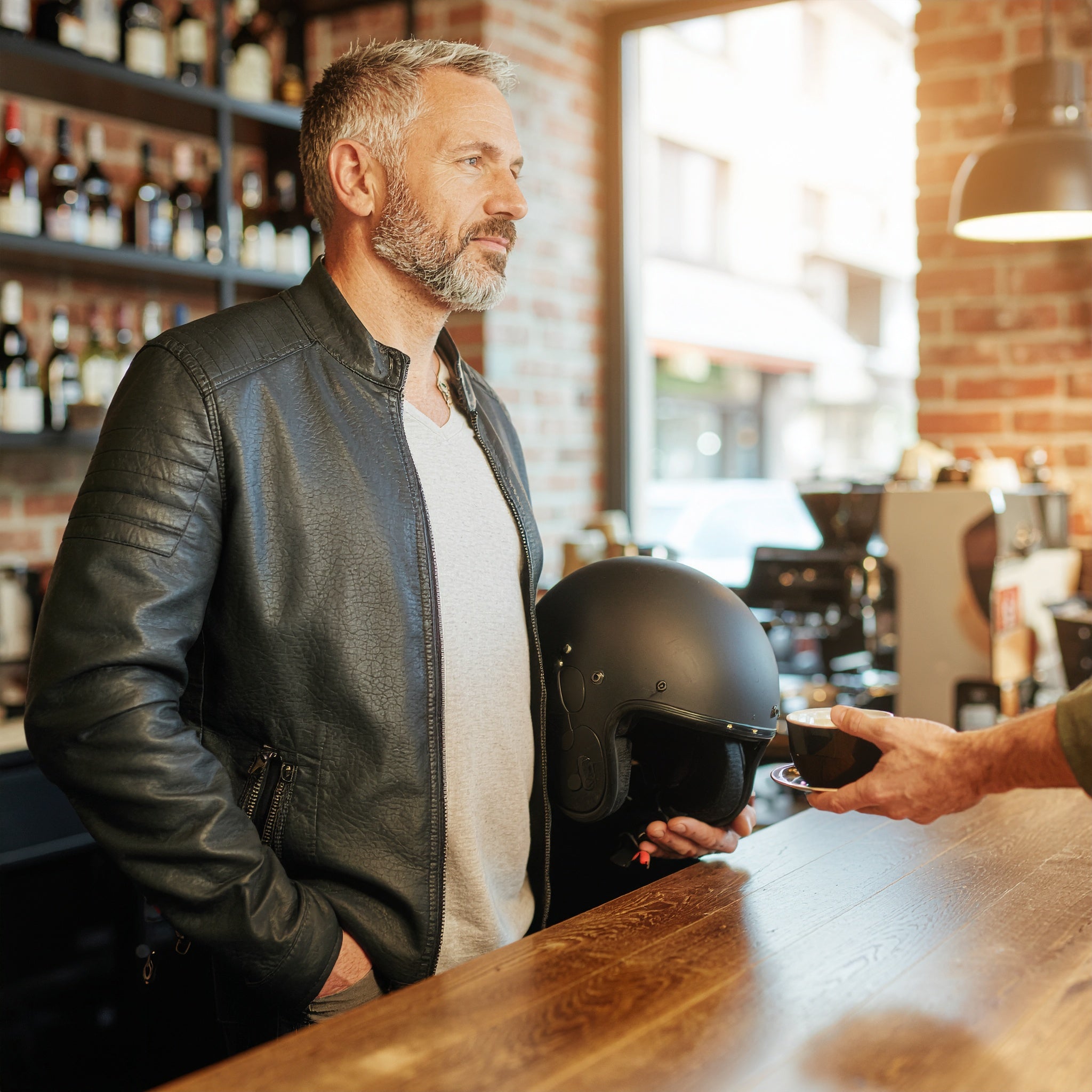 A confident man in a black leather jacket stands at a coffee shop counter holding a matte black motorcycle helmet while receiving a cup of coffee, sunlight filtering through the window, capturing effortless masculine energy and calm confidence.