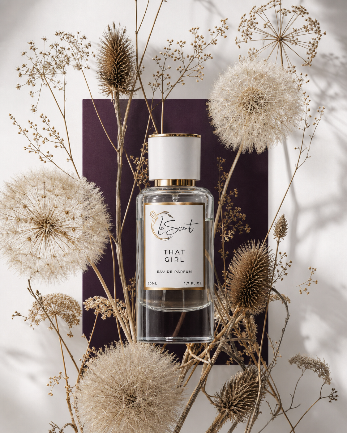A clear glass perfume bottle labeled 'LeScent THAT GIRL' with a white cap and gold trim, styled on a dark purple rectangular card surrounded by dried flowers and botanicals against a white background.