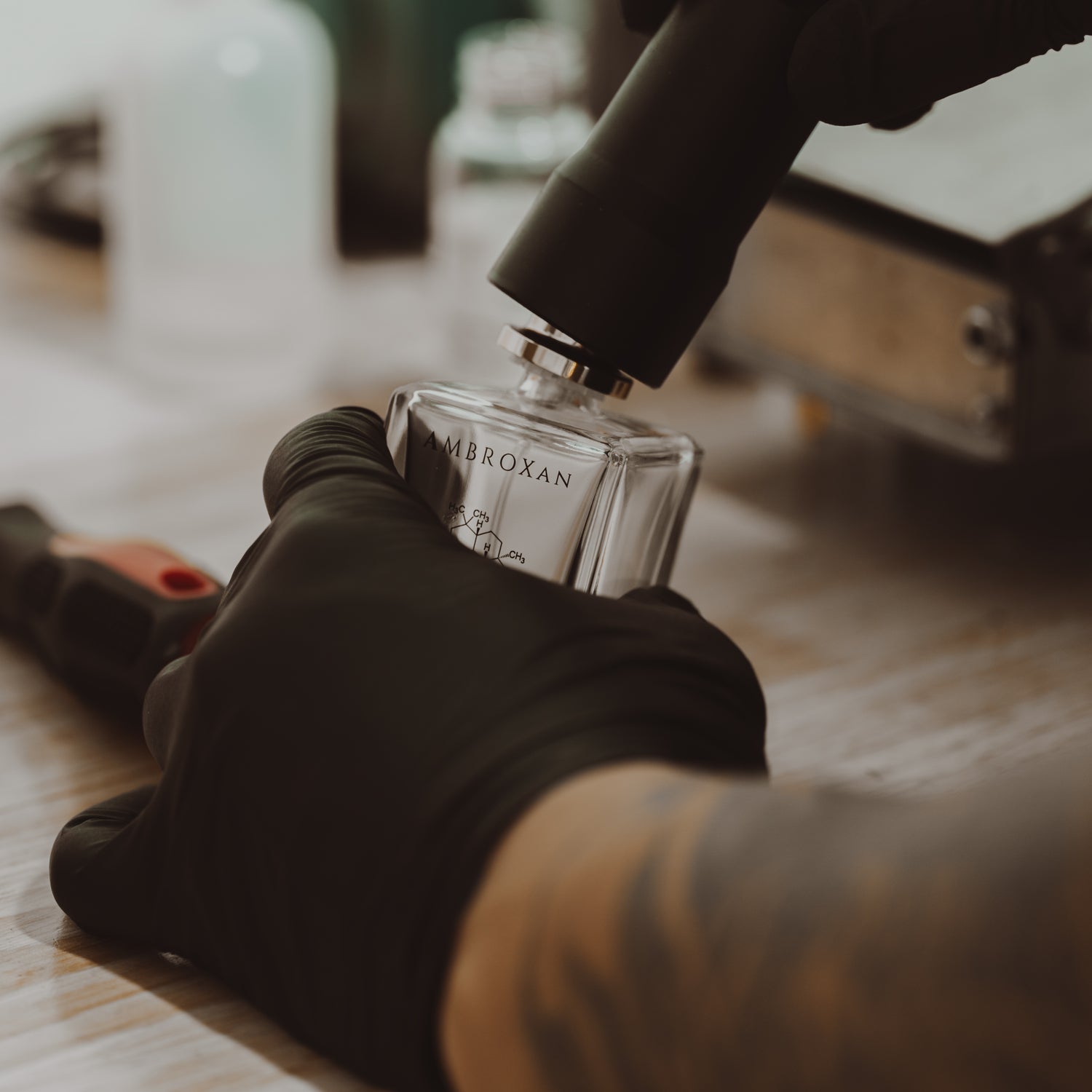 A close-up of a worker in black gloves capping a bottle of LeScent Ambroxan perfume on a wooden lab surface.