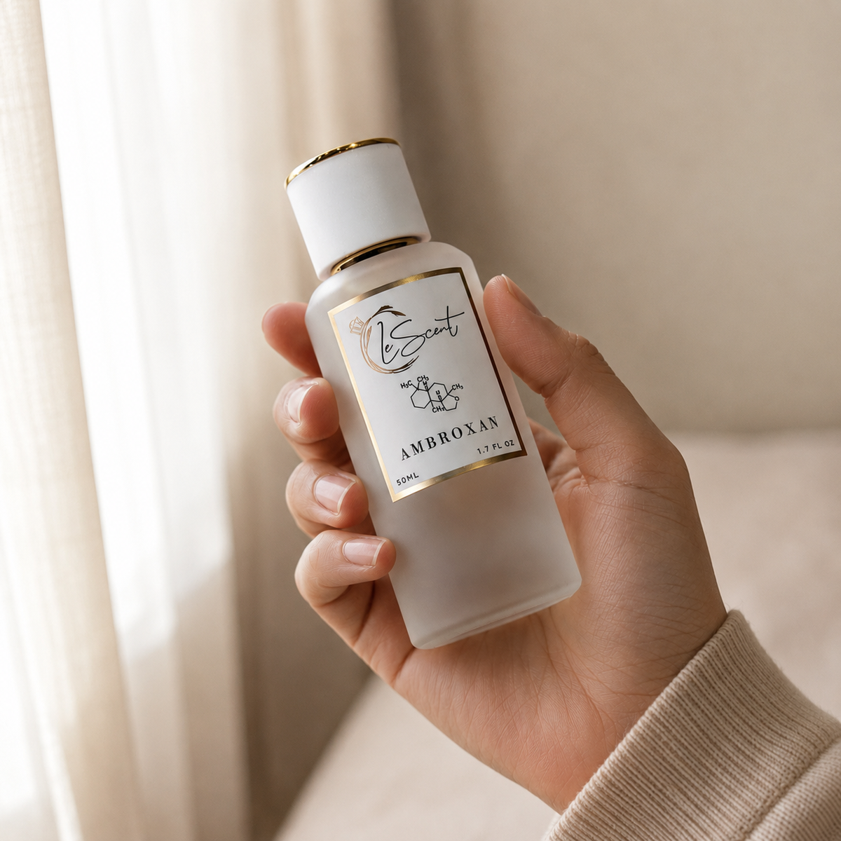 A hand holding a frosted glass perfume bottle with a white cap and gold trim, labeled 'Bscint Ambroxat 100ml'. The background is a softly lit, warm beige interior with sheer curtains.
