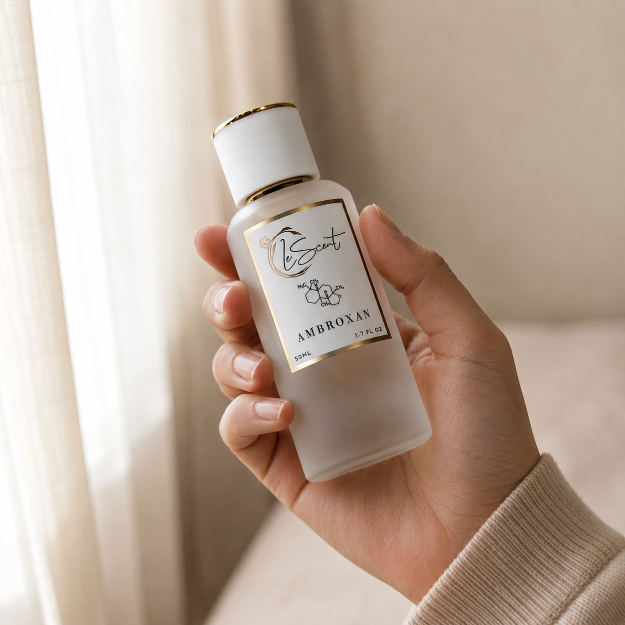 A hand holding a frosted glass perfume bottle with a white cap and gold trim, labeled 'Bscint Ambroxat 100ml'. The background is a softly lit, warm beige interior with sheer curtains.