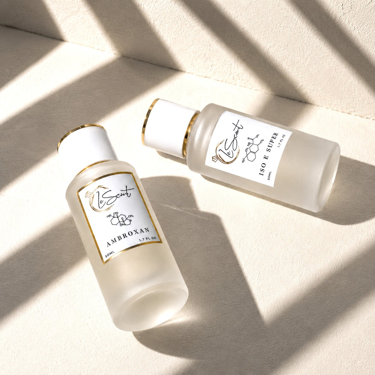 Two frosted glass perfume bottles with white caps and gold trim, labeled 'LScent' and 'AMBROXAN' and 'NO E SPICE', arranged diagonally on a textured beige surface with soft window shadow patterns.