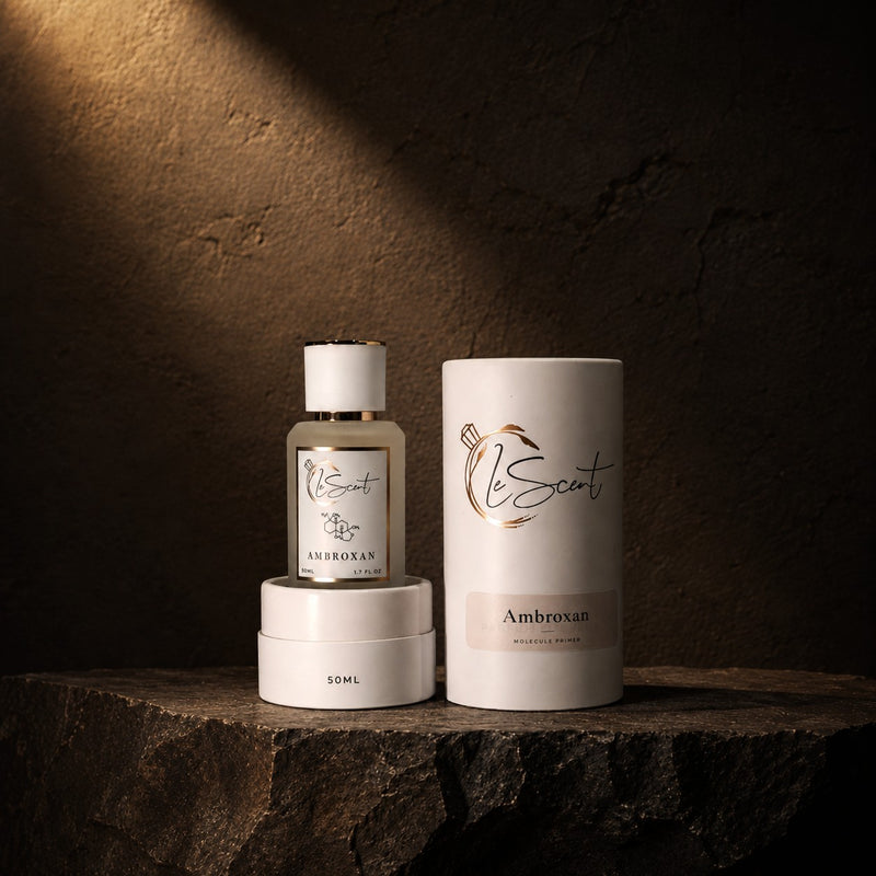 A perfume bottle and its cylindrical packaging box, both in white with gold and black detailing, displayed on a dark stone surface against a textured dark brown background with dramatic lighting.