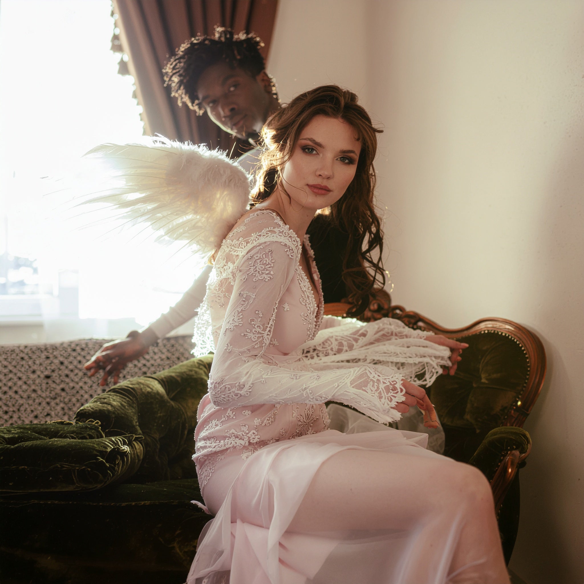 A woman in a sheer, lace gown with angel wings seated on a green velvet sofa, with a man standing in the background, set in a bright, romantic interior.