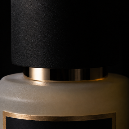 Close-up of a luxury bottle with a matte black textured cap, a polished gold collar, and a frosted beige glass body with a gold-trimmed black label.