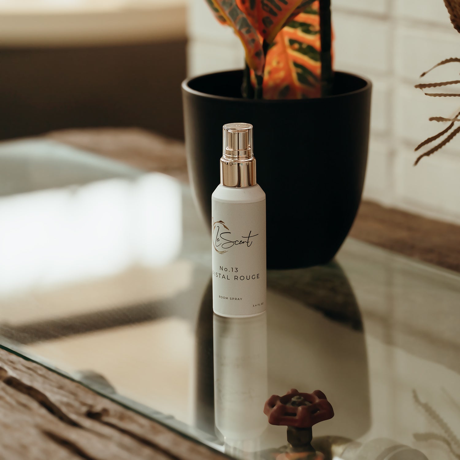 LeScent Crystal Rouge room spray bottle with gold nozzle on glass table beside indoor plant, warm natural lighting interior shot