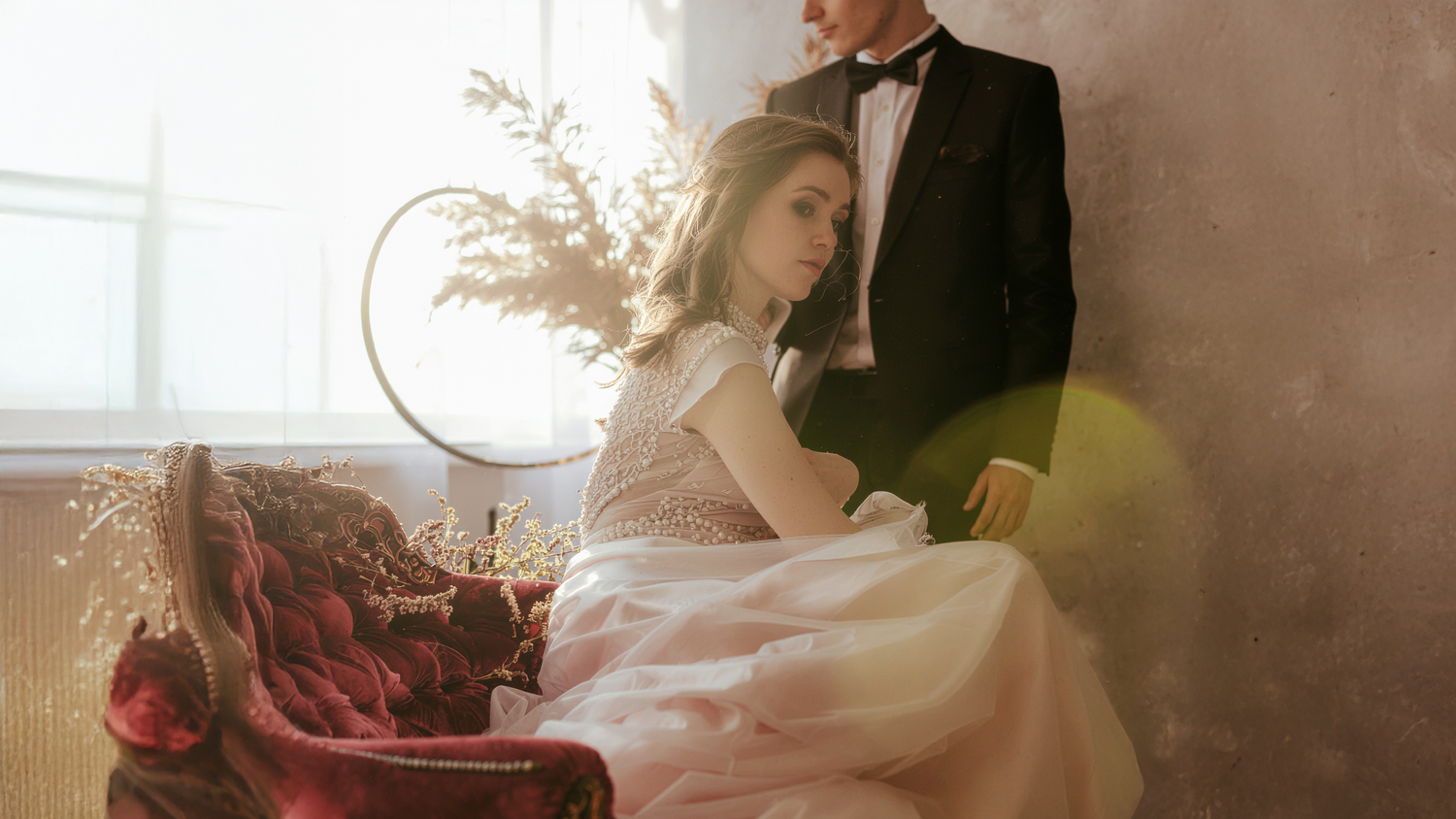 A wide shot of a woman in an elegant, pale pink gown seated on a deep red velvet chair, with a man in a tuxedo standing nearby.