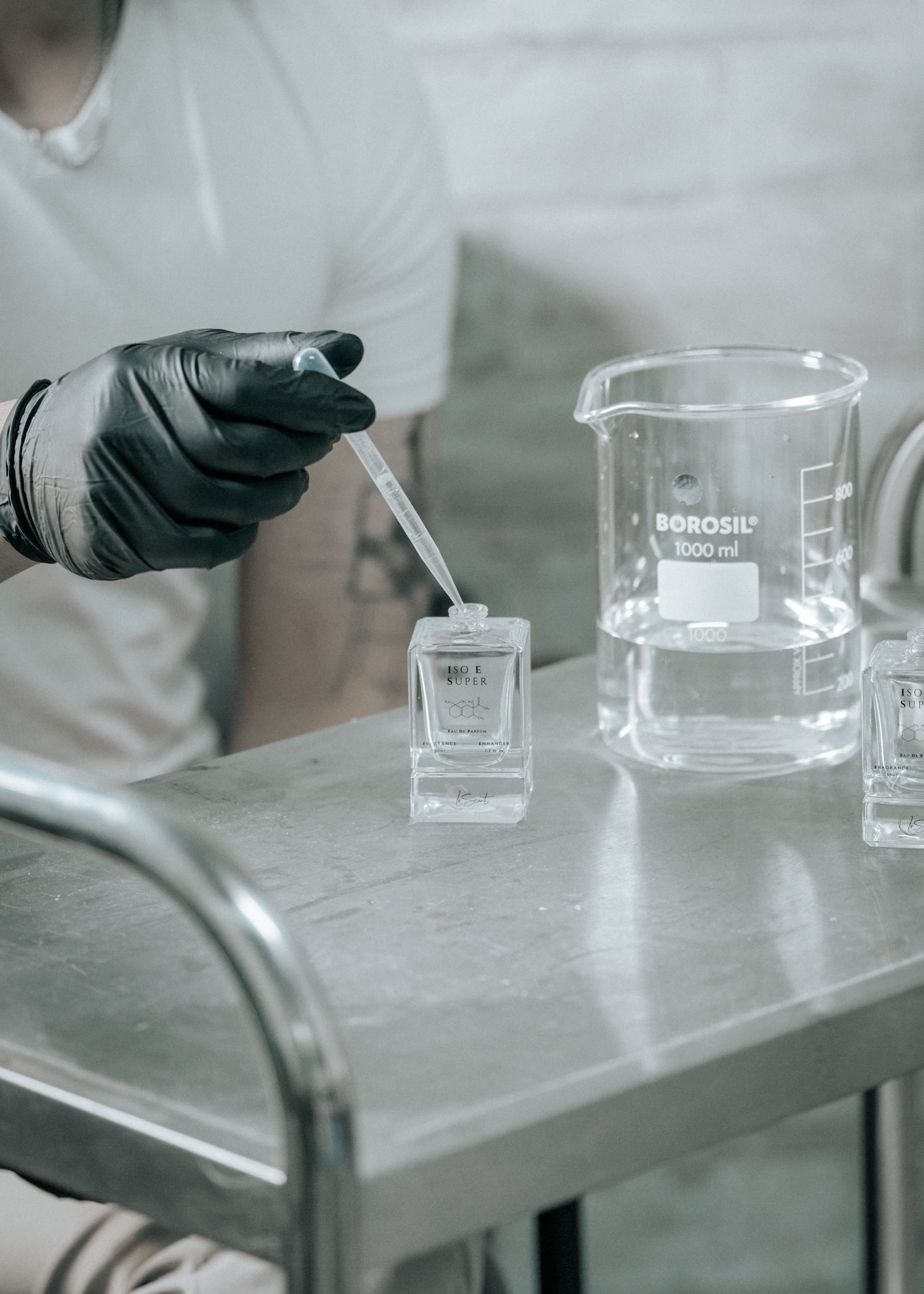 A person wearing a black glove uses a glass dropper to fill a LeScent ISO E SUPER bottle, with a large glass beaker on a stainless steel lab table.