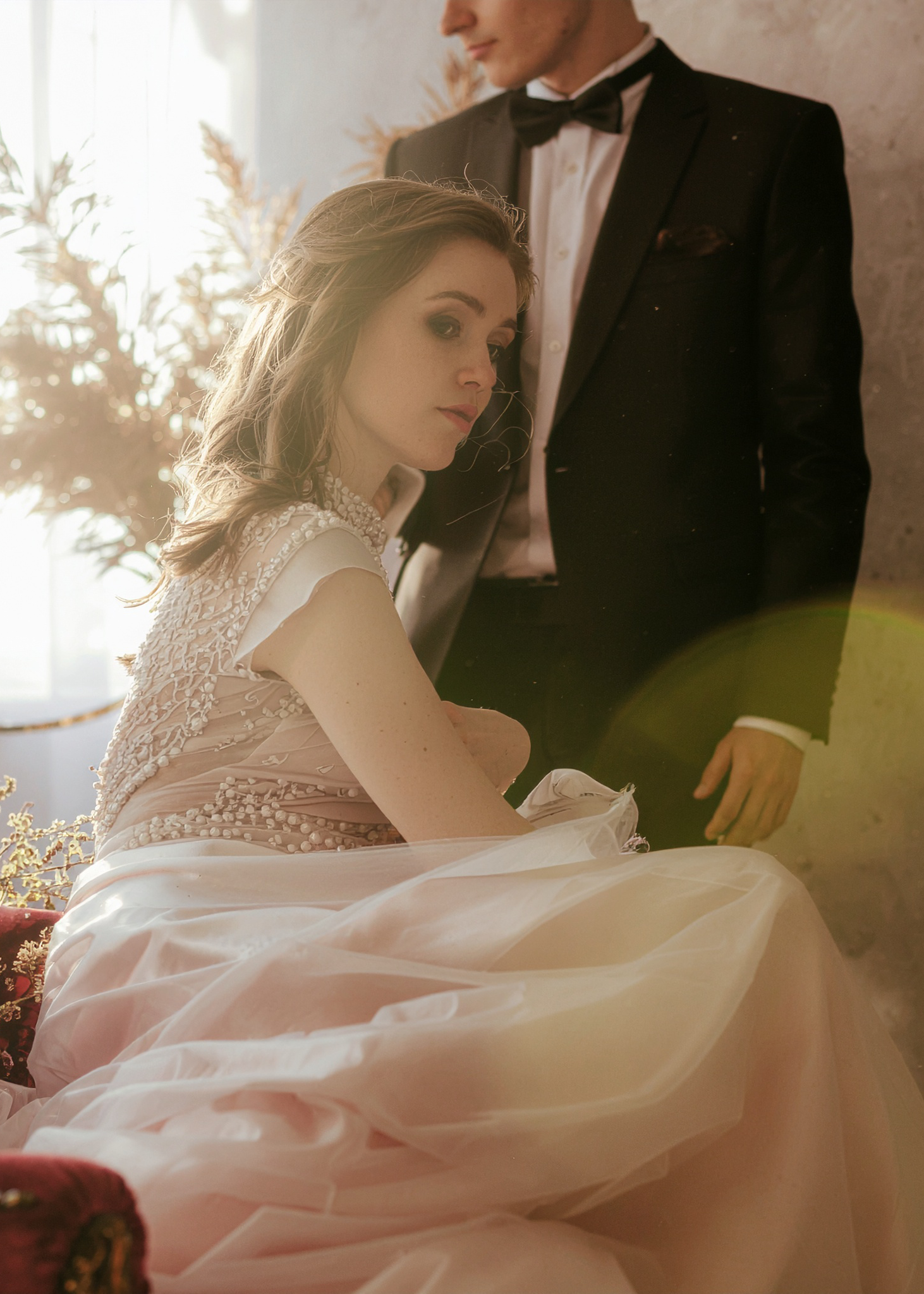 A woman in an elegant, beaded gown seated in a velvet chair, with a man in a black tuxedo standing behind her, symbolizing sophisticated romance and luxury.