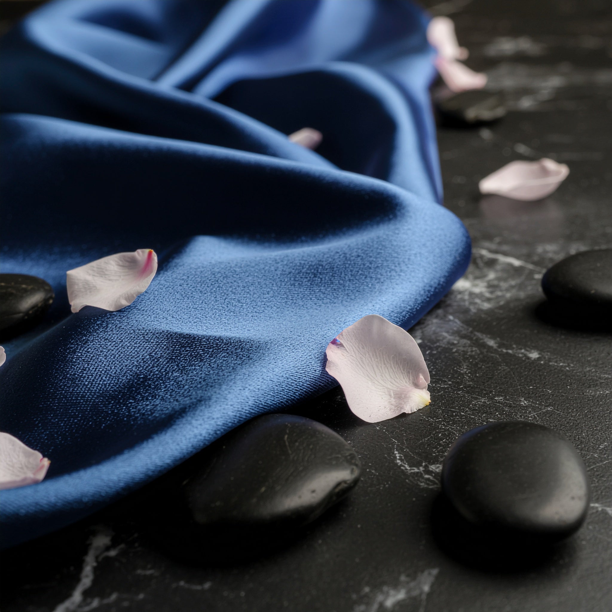 A luxurious composition of dark blue satin silk draped over a black marble surface, accented by pink rose petals and smooth black river stones.
