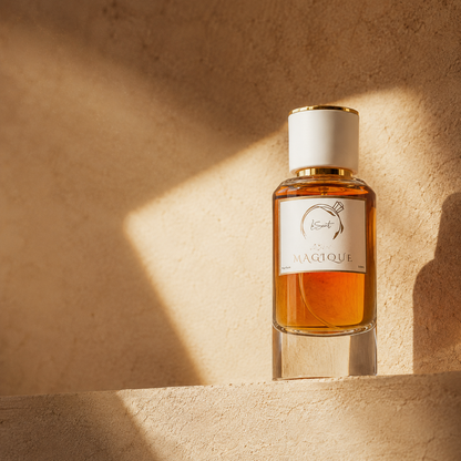 A bottle of LeScent Magique perfume with amber liquid, a white cap, and a white label, set on a textured beige stone surface with dramatic warm sunlight and shadows.