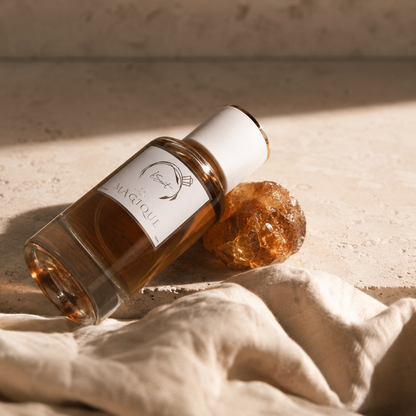 A clear glass bottle with amber liquid and a white cap lies on a beige stone surface next to a rough amber crystal, with draped cream fabric in the foreground and a soft shadow cast across the scene.