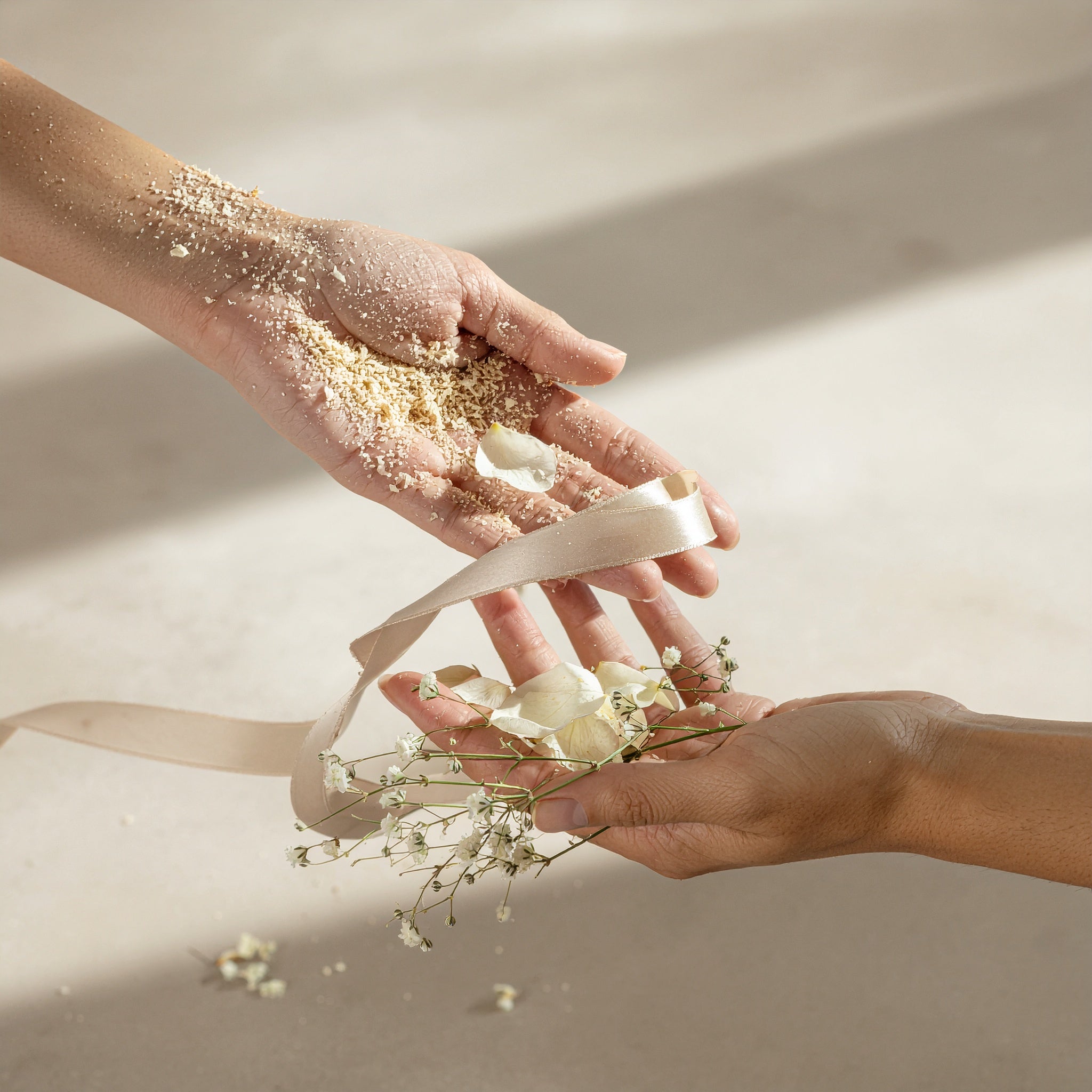 Two hands gently exchanging elements: one holding sand and petals, the other holding baby's breath flowers and a satin ribbon, symbolizing delicate natural ingredients.
