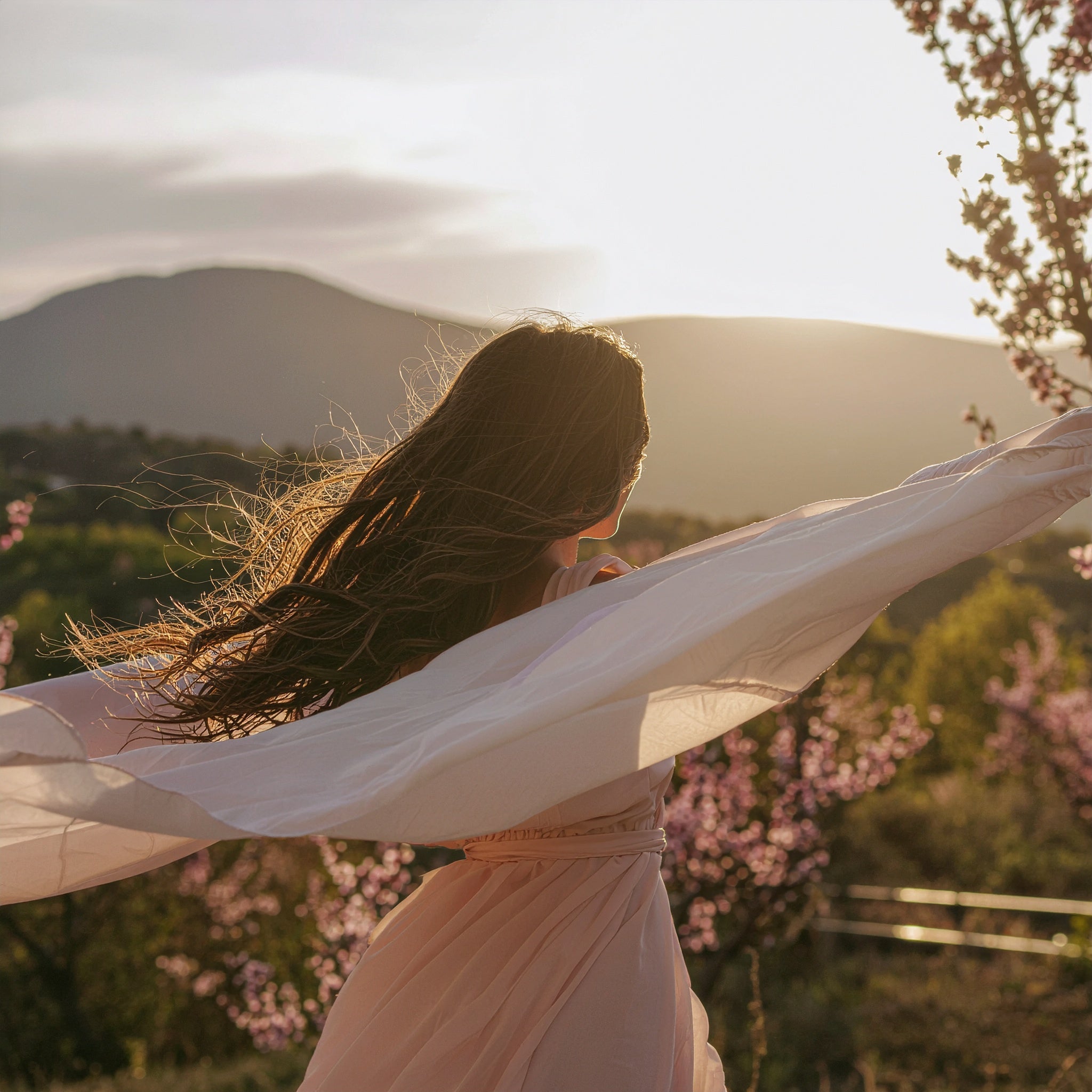 A woman standing in a field of pink blossoms with her back to the camera, holding a pale pink scarf that billows in the wind against a mountain sunset backdrop.
