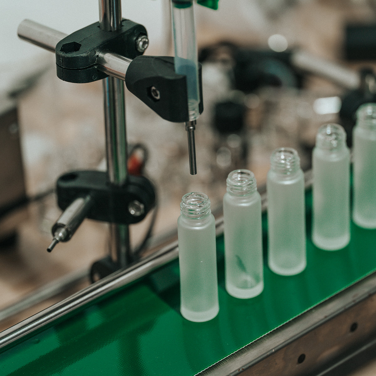 A close-up of an automated bottling machine filling a row of frosted glass perfume oil bottles on a green conveyor belt.