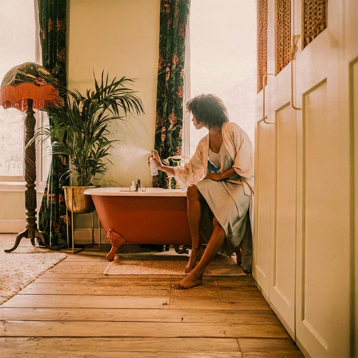 Woman spraying LeScent room spray beside a freestanding bathtub in a warm, elegant bathroom, home fragrance spray and room freshener lifestyle image.