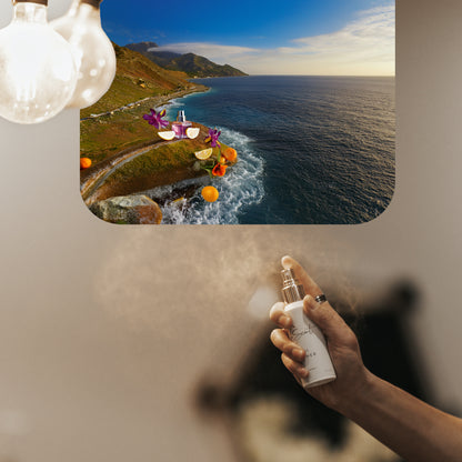 Hand spraying LeScent Sweet Aqua room fragrance freshener mist, with an ingredient panel showing oranges, sliced lime, purple flowers, and green leaves arranged on a coastal cliff overlooking blue ocean waves, inspired by Creed Millésime Impérial.