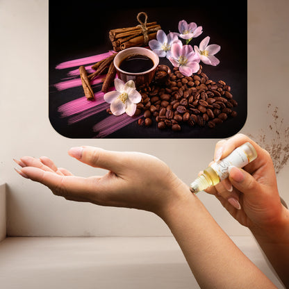 Applying LeScent Vanille Café roll-on perfume oil beside roasted coffee beans, cinnamon sticks, and pink floral accents, inspired by Yves Saint Laurent Black Opium.